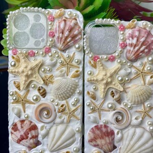May include: Two white phone cases decorated with seashells, pearls, and pink flowers. The cases have a cutout for the camera lens.