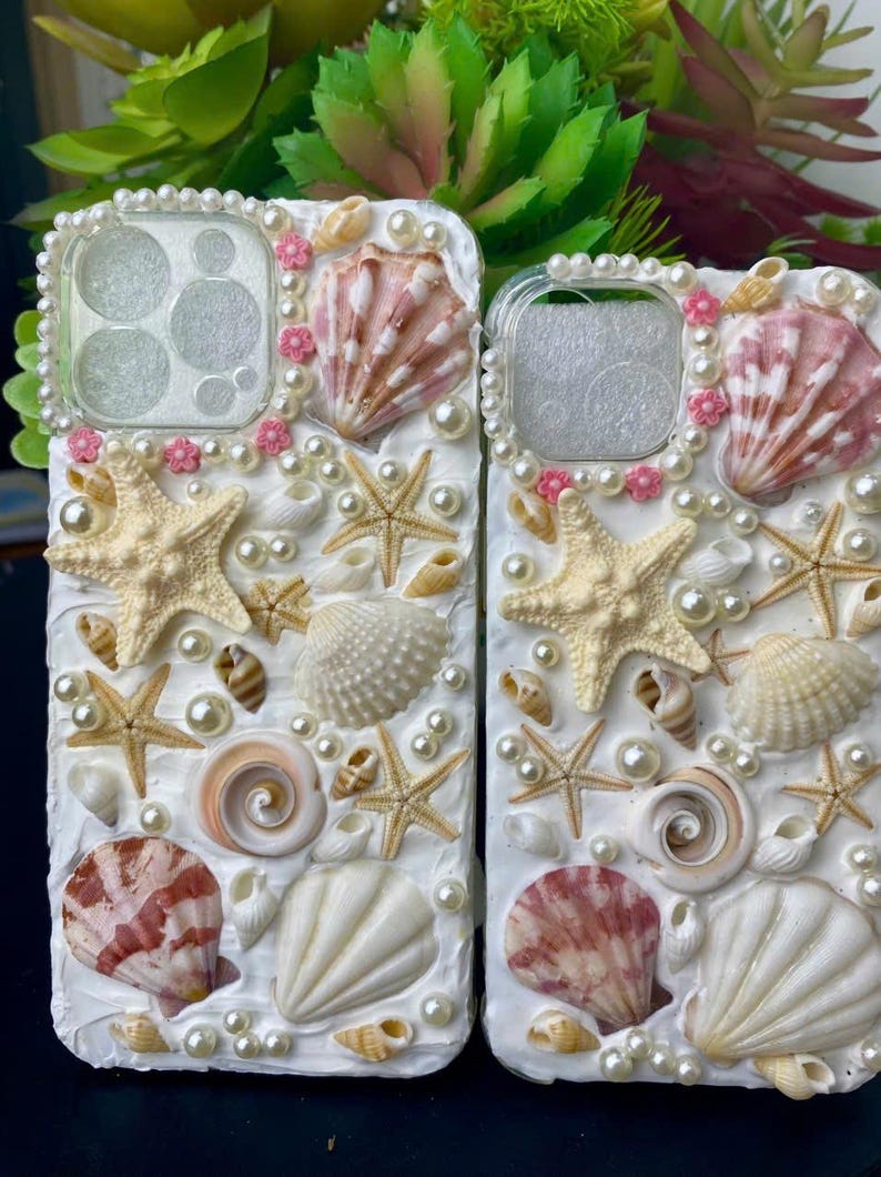 May include: Two white phone cases decorated with seashells, pearls, and pink flowers. The cases have a cutout for the camera lens.