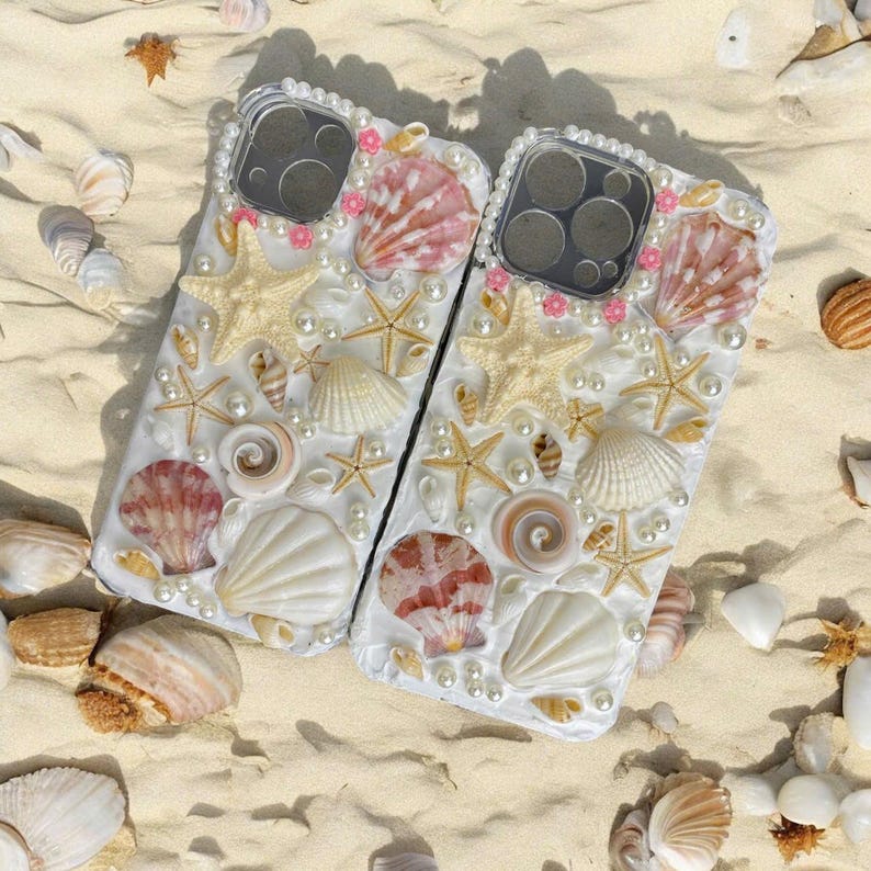 May include: Two white phone cases decorated with seashells, pearls, and starfish. The cases have a clear back and a white front. The shells are in various sizes and colors, including pink, white, and brown.