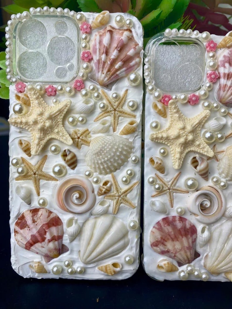 May include: A white phone case decorated with seashells, starfish, and pearls. The case has a clear back panel and a white border.
