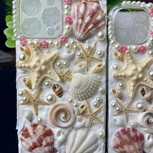May include: A white phone case decorated with seashells, pearls, and starfish. The case has a clear back panel and a white front panel. The seashells are in various sizes and shapes, and the pearls are white. The starfish are beige.
