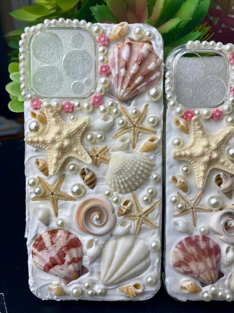 May include: A white phone case decorated with seashells, pearls, and starfish. The case has a clear back panel and a white front panel. The seashells are in various sizes and shapes, and the pearls are white. The starfish are beige.