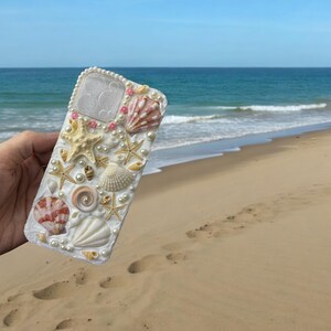 May include: White iPhone case decorated with seashells, pearls, and pink accents. The case is designed to look like a beach scene.