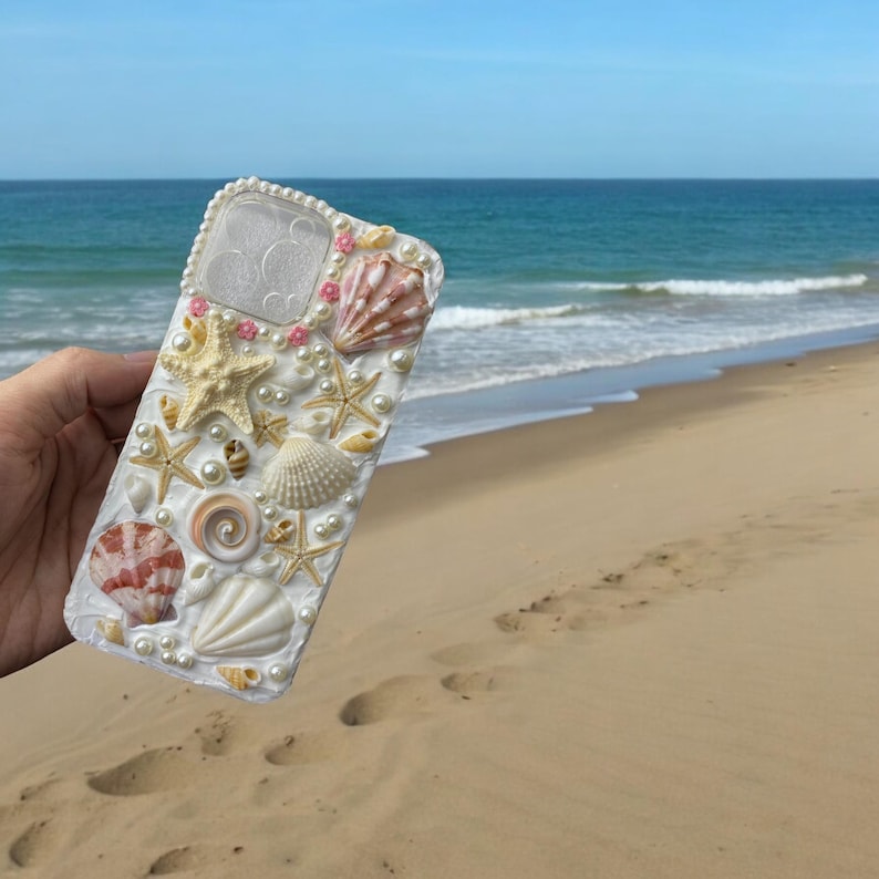 May include: White iPhone case decorated with seashells, pearls, and pink accents. The case is designed to look like a beach scene.