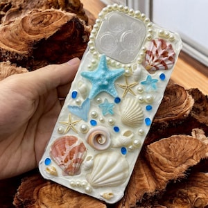 May include: A white phone case decorated with seashells, pearls, and starfish. The case has a blue and white color scheme and features a variety of different seashells, including clams, conch shells, and scallop shells.