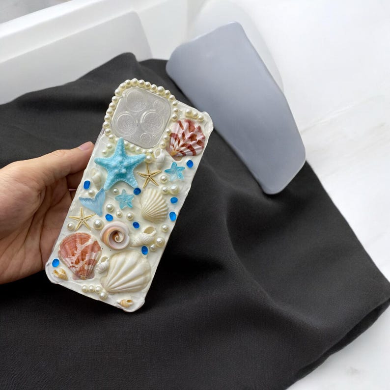 May include: White phone case decorated with seashells, pearls, and blue starfish.