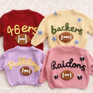 May include: Four long-sleeved knit jumpers in red, cream, lavender, and pink. Each jumper features a central brown football appliqu&eacute; with white stitching. The red jumper says 46ers, the cream says backers with blue flowers, the lavender says Pulldogs, and the pink says Raidons with black butterflies and the letters PJ on the sleeve.
