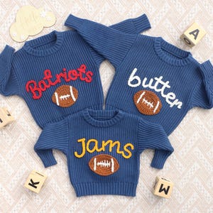 May include: Three blue long-sleeved knit sweaters featuring embroidered text and a brown football applique on the front. The top left sweater says Batriots in red, the top right says butter in white, and the bottom sweater says jams in yellow.