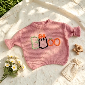 May include: A pink long-sleeve knit sweater featuring a crew neckline and ribbed cuffs. The front displays the word BOO embroidered in yarn, with the letter B in light green, a ghost wearing an orange bow, and two orange circles for the Os.