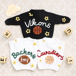 May include: Three long-sleeve knit jumpers featuring textured yarn appliques. The top black jumper displays the text Vikons with a basketball. The bottom white jumpers feature the text Backers with a football and Cavadiers with a basketball.
