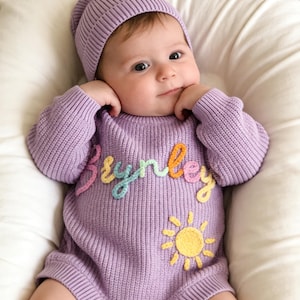 Puede incluir: Un mameluco de punto de manga larga en color violeta claro con un gorro a juego. El mameluco presenta el nombre Brynley bordado en hilo multicolor en el pecho y una aplicaci&oacute;n de sol amarillo debajo del nombre. La prenda tiene tres botones marrones.