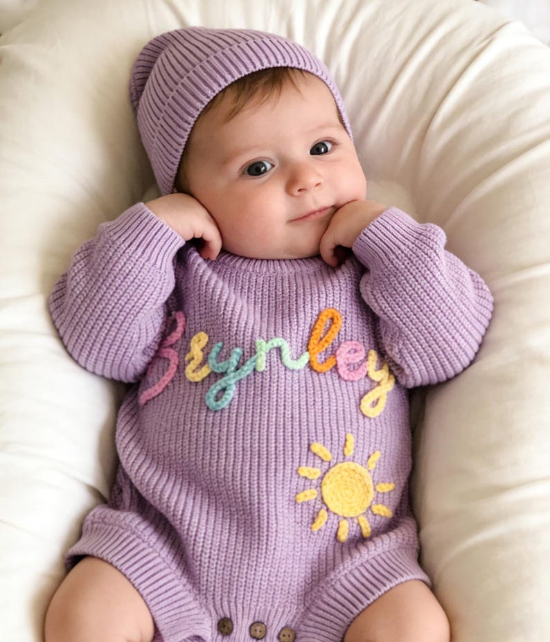 Puede incluir: Un mameluco de punto de manga larga en color violeta claro con un gorro a juego. El mameluco presenta el nombre Brynley bordado en hilo multicolor en el pecho y una aplicaci&oacute;n de sol amarillo debajo del nombre. La prenda tiene tres botones marrones.