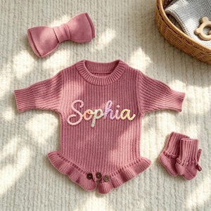Puede incluir: Un pelele de punto rosa empolvado con textura acanalada, dobladillo con volantes y tres botones en la parte inferior. El nombre Sophia est&aacute; bordado con hilo multicolor en el pecho. Incluye un lazo de punto a juego y dos patucos peque&ntilde;os.