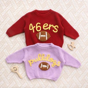 May include: Two long-sleeved knit sweaters. The top sweater is dark red with yellow text reading 46ers and a brown football applique. The bottom sweater is light purple with yellow text reading bulldogs and a brown football applique.