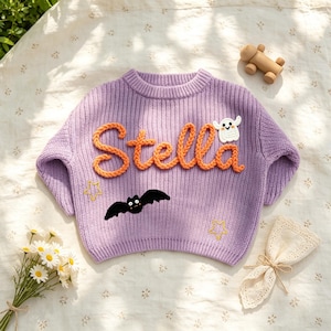 May include: A lavender long-sleeve knit sweater featuring the name Stella in raised orange yarn lettering. The sweater is decorated with a white ghost appliqu&eacute;, a black bat appliqu&eacute;, and two yellow embroidered stars on the front.
