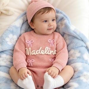 Puede incluir: Un pelele de punto rosa de manga larga con el nombre Madeline en letras blancas, decorado con peque&ntilde;os lazos bordados en rosa. El conjunto incluye un gorro de punto rosa a juego y calcetines blancos sobre una manta azul claro con flores.