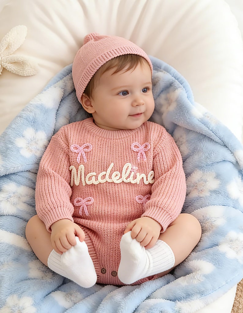 Puede incluir: Un pelele de punto rosa de manga larga con el nombre Madeline en letras blancas, decorado con peque&ntilde;os lazos bordados en rosa. El conjunto incluye un gorro de punto rosa a juego y calcetines blancos sobre una manta azul claro con flores.