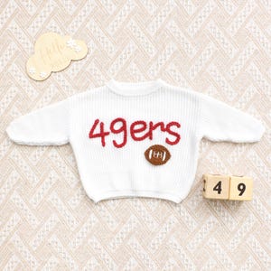 May include: White knit sweater with the word "49ers" in red and a brown football applique. The sweater is displayed on a patterned surface, with wooden blocks showing the numbers 1 and 4, and 9.