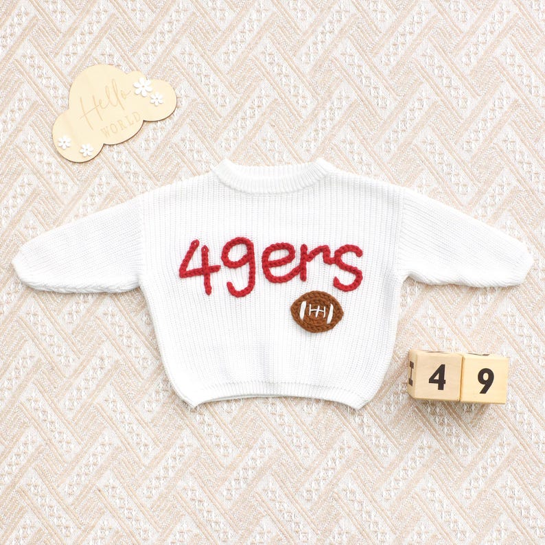 May include: White knit sweater with the word "49ers" in red and a brown football applique. The sweater is displayed on a patterned surface, with wooden blocks showing the numbers 1 and 4, and 9.