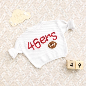 May include: A white long-sleeve knit jumper featuring the red embroidered text 46ers and a brown football graphic on the front. A wooden cloud-shaped sign with the text Hello World and two wooden blocks with the numbers 4 and 9 are present.