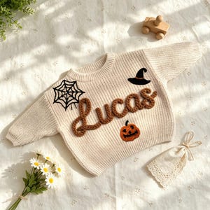 May include: A cream-colored long-sleeve knit sweater featuring the name Lucas in brown yarn, a black embroidered spider web, a black witch hat, and an orange jack-o'-lantern pumpkin patch on the front chest area of the garment.