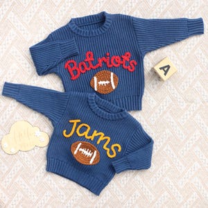 May include: Two blue long-sleeve knit jumpers featuring textured appliques. The top jumper has the red text "Batriots" above a brown football. The bottom jumper has the yellow text "Jams" above a brown football. Both have ribbed cuffs.