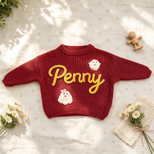 May include: A deep red long-sleeve knit sweater featuring the name Penny embroidered in yellow yarn across the center. The sweater is decorated with two small white crocheted ghost-like figures, one with a red bow and one with a pink bow.