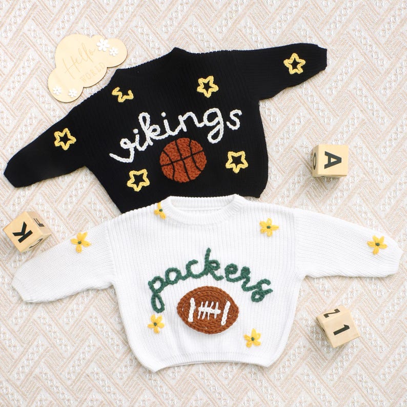 May include: Two knitted sweaters, one black and one white, with sports-themed designs. The black sweater features the word "vikings" and a basketball, while the white sweater says "packers" with a football. Both sweaters have star and flower accents.