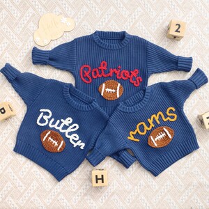 May include: Three navy blue knit sweaters with crew necks. Each sweater features a team name in a contrasting color and a football applique. The sweaters are personalized with the names "Patriots", "Butler", and "rams".