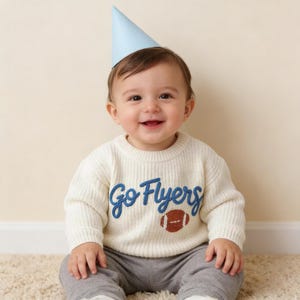 May include: A light blue party hat worn with a cream-coloured long-sleeve knit sweater featuring the blue embroidered text Go Flyers and a brown football graphic, paired with grey trousers, creating a festive and casual outfit for a young child.