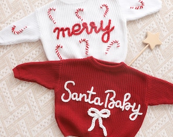 Pode incluir: Duas camisolas de Natal. Uma é branca com detalhes de bengalas vermelhas e a palavra "merry" em vermelho. A outra é vermelha com as palavras "Santa Baby" em branco e um laço branco.
