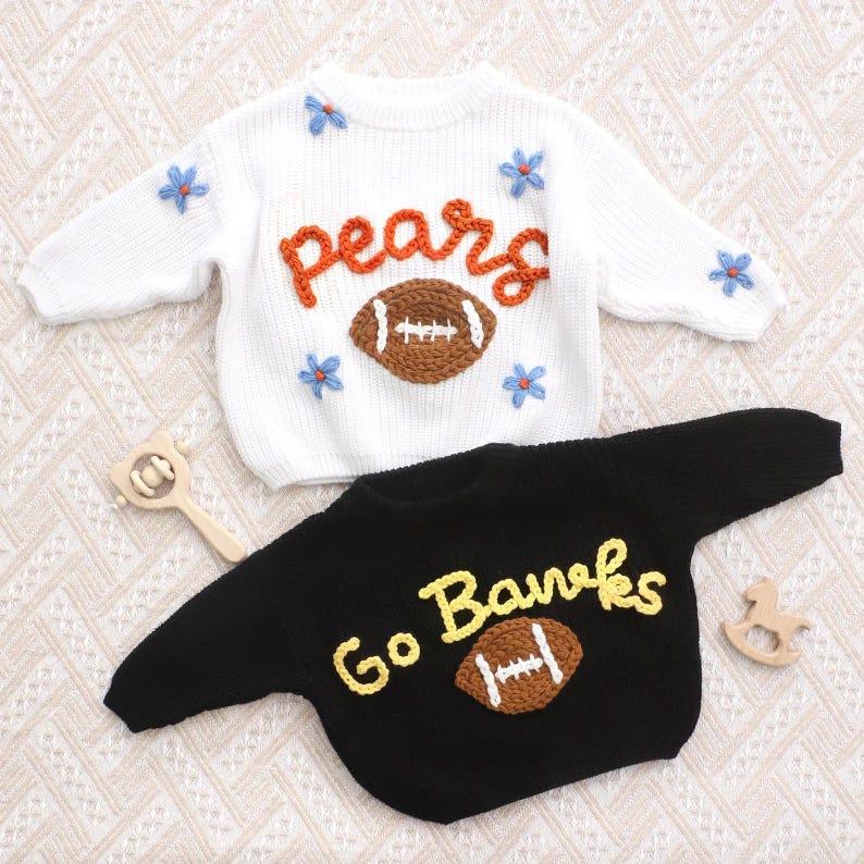 May include: A white long-sleeve sweater with orange text reading "pears" and a brown football graphic, accented with small blue flowers. A black long-sleeve sweater with yellow text reading "Go Banks" and a brown football graphic.