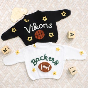 May include: A black knit jumper with white text reading Vikons, a brown basketball graphic, and yellow star accents, paired with a white knit jumper featuring green text reading Backers, a brown football graphic, and yellow flower accents.