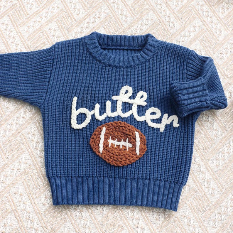 May include: A blue long-sleeve knit sweater featuring the word butter in white yarn lettering above a brown football graphic with white stitching details. The garment has a ribbed crew neckline, ribbed cuffs, and a ribbed hemline design.