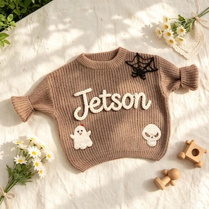 May include: A tan ribbed knit sweater featuring the name Jetson in white yarn lettering. The sweater is decorated with a black yarn spiderweb, a white ghost applique, and a white skull applique. It has long sleeves with flared cuffs.