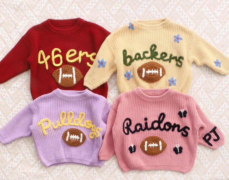 May include: Four long-sleeved knit jumpers in red, cream, lavender, and pink. Each jumper features a central brown football appliqu&eacute; with white stitching. The red jumper says 46ers, the cream says backers with blue flowers, the lavender says Pulldogs, and the pink says Raidons with black butterflies and the letters PJ on the sleeve.