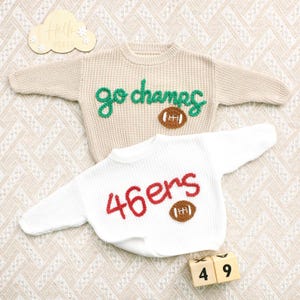 May include: A beige long-sleeve knit jumper with green embroidered text reading go champs and a brown football applique, and a white long-sleeve knit jumper with red embroidered text reading 46ers and a brown football applique.