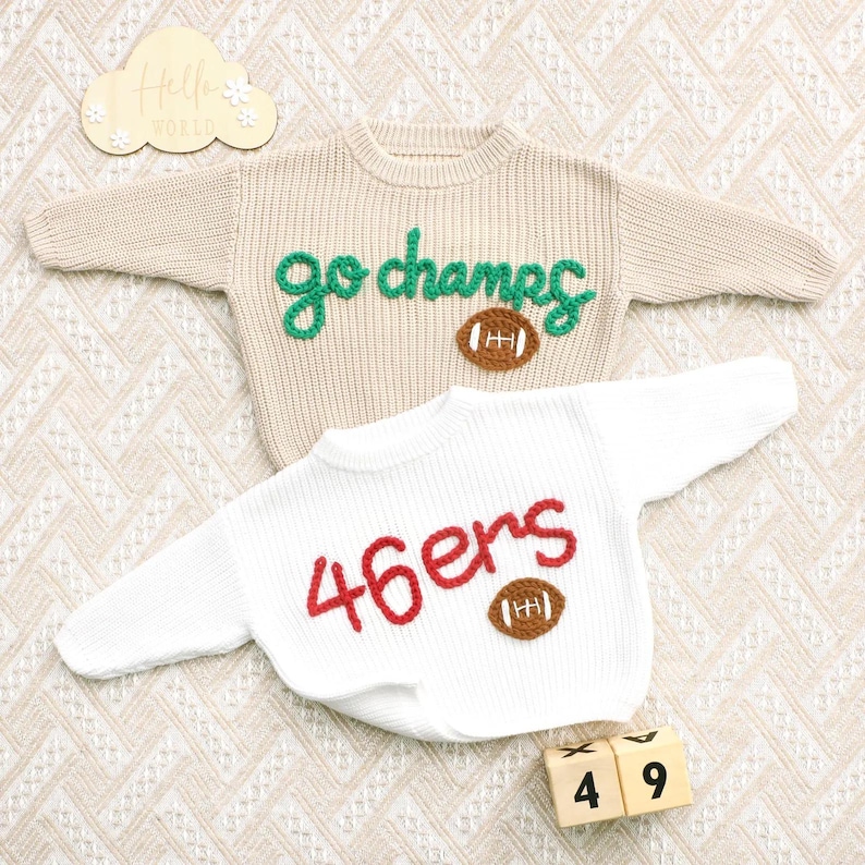 May include: A beige long-sleeve knit jumper with green embroidered text reading go champs and a brown football applique, and a white long-sleeve knit jumper with red embroidered text reading 46ers and a brown football applique.