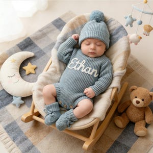 Puede incluir: Un pelele de punto de manga larga en color azul polvoriento con el nombre Ethan bordado en letra cursiva blanca en el pecho, junto con un gorro a juego con pomp&oacute;n y patucos de punto con lazos, usado por un beb&eacute; durmiendo.