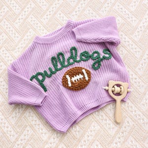 May include: A light purple long-sleeve knit jumper featuring the word "Bulldogs" embroidered in dark green yarn across the chest, accompanied by a brown knit football graphic and a light-coloured wooden rattle with a bear-shaped handle.