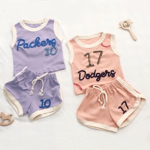 Pode incluir: Dois conjuntos de roupa desportiva para crianças. Um conjunto é lavanda com "Packers 10" e o outro é pêssego com "Dodgers 17". Cada conjunto inclui um top sem mangas e calções a condizer com acabamento branco e cordão.