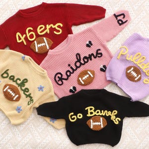 May include: Five long-sleeved knit jumpers in red, cream, pink, lavender, and black. Each jumper features a brown crocheted football appliqu&eacute; and embroidered text including 46ers, backers, Raidons, Pulldogs, and Go Banks, with some floral accents.