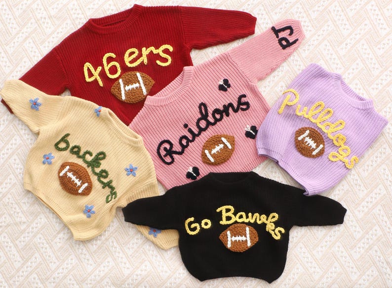 May include: Five long-sleeved knit jumpers in red, cream, pink, lavender, and black. Each jumper features a brown crocheted football appliqu&eacute; and embroidered text including 46ers, backers, Raidons, Pulldogs, and Go Banks, with some floral accents.