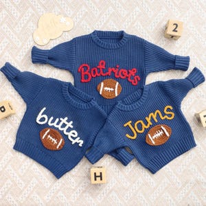 May include: Three navy blue long-sleeve knit jumpers with ribbed collars and cuffs. The top jumper features red text reading "Batriots" and a brown football. The bottom left jumper has white text reading "butter" and the bottom right has yellow text reading "Jams", both with brown footballs.