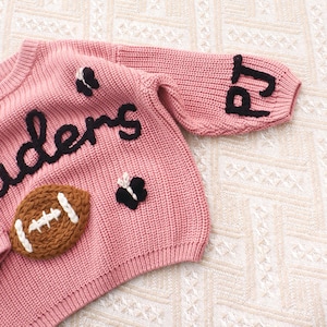 May include: A dusty pink knit jumper featuring black embroidered text that reads 'ders' and 'pj' on the sleeve. The jumper is decorated with a brown football applique with white stitching and two small black and white butterfly appliques.