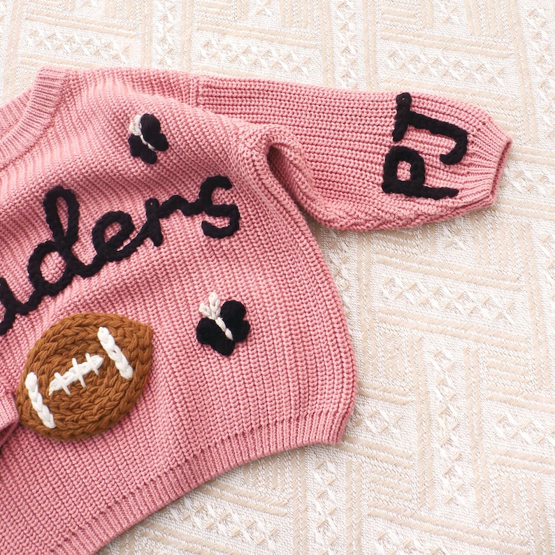 May include: A dusty pink knit jumper featuring black embroidered text that reads 'ders' and 'pj' on the sleeve. The jumper is decorated with a brown football applique with white stitching and two small black and white butterfly appliques.