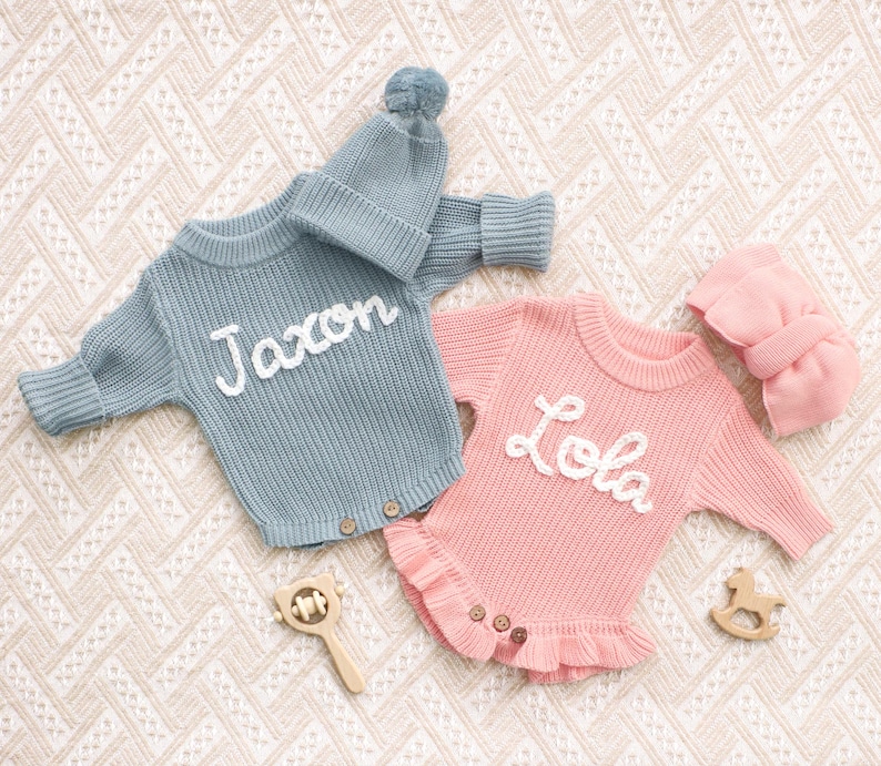Puede incluir: Un pelele de punto azul de manga larga con el nombre Jaxon bordado en blanco, junto a un gorro con pomp&oacute;n a juego, y un pelele de punto rosa de manga larga con el nombre Lola bordado en blanco, que presenta un dobladillo con volantes.