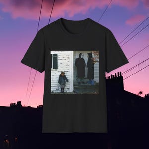 May include: Black t-shirt featuring a photograph of three figures in front of a white house. One figure wears a skull mask and long black coat. A child in a plaid skirt and jacket stands to the left. The sky is a gradient of pink and purple.