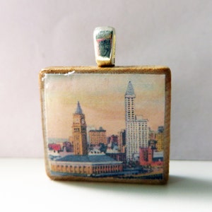 May include: A wooden pendant with a vintage postcard image of a city skyline. The postcard shows a tall building with a pointed roof and several other buildings in the background. The pendant has a silver bail.