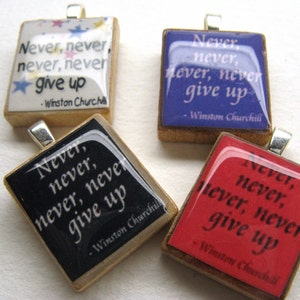Puede incluir: Cuatro colgantes cuadrados de madera con un aro de plata. Cada colgante tiene un fondo negro con texto blanco que dice "Never, never, never, never give up - Winston Churchill". Los colgantes son de diferentes colores: blanco con estrellas, negro, morado y rojo.